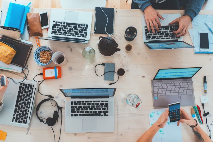 Vista dall'alto di un tavolo con computer, una persona che scrive al pc e un’altra al telefono, senza volti visibili